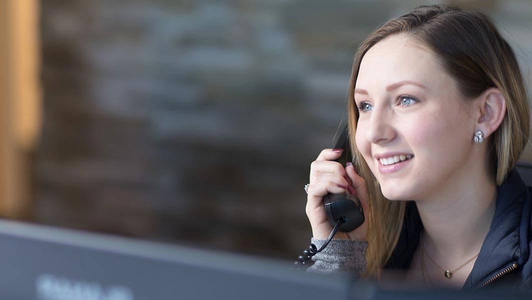 A friendly woman speaking on a telephone, smiling while seated at a desk, with a blurred background and warm lighting creating a professional yet approachable setting.
